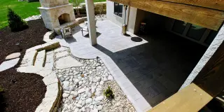 Paver patio beneath a covered deck with an outdoor fireplace and a river rock drainage bed